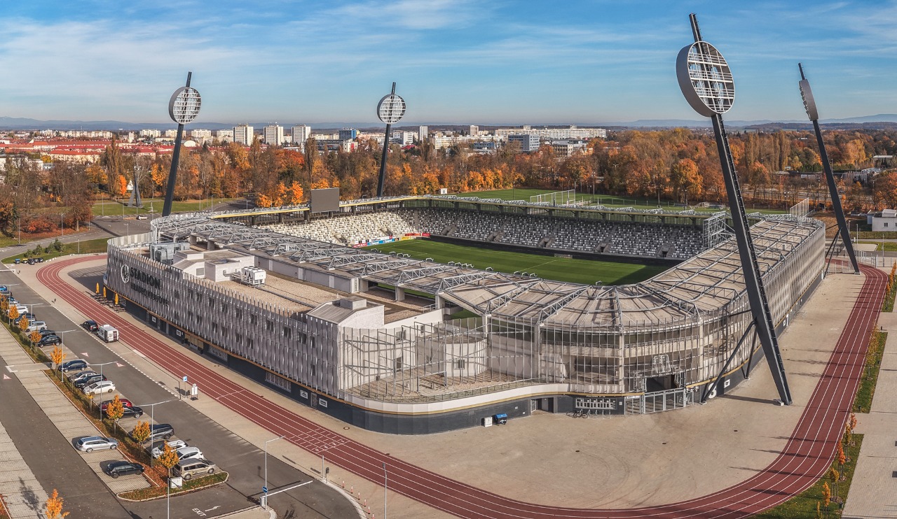 Aerial view of the new Malšovice Stadium in Hradec Králové, officially known as the Všesportovní stadion. The modern football stadium was completed in 2023 on the site of the original 1960s venue. It has a capacity of approximately 9,300 spectators and serves as the home ground of FC Hradec Králové. The image shows the entire arena with its four distinctive floodlight pylons, surrounding parking areas, and the cityscape in autumn colors.