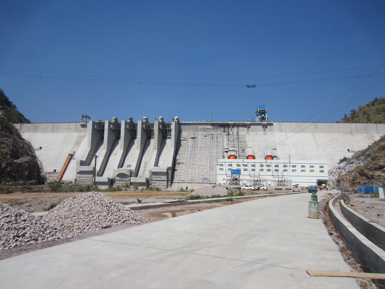Bui Dam, Ghana in November 2013 shortly before official opening. Looking upstream, approximately north-east. Powerhouse at right, with three orange penstocks visible above the roof (one for each generator). Spillway has five bays. The penstock for the future house unit is visible at the left. The dam crest is about 75 metres above the powerhouse main floor. Powerhouse paved road under construction visible in foreground.